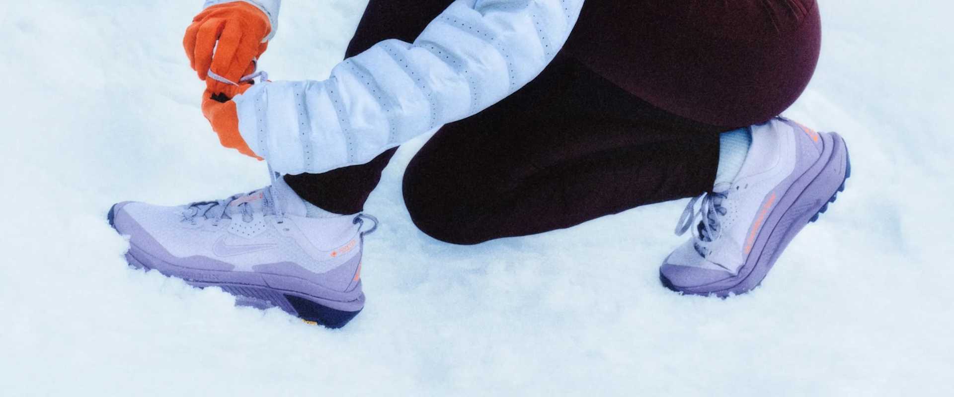 Nahaufnahme von Händen in orangen Handschuhen, die einen Trailschuh im Schnee schnüren