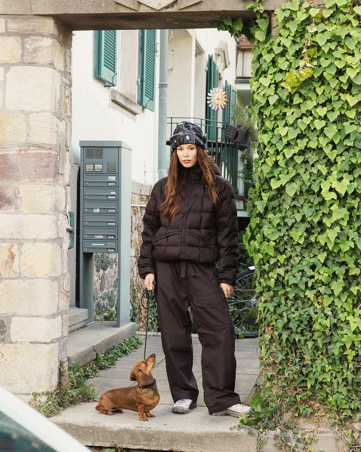 Frau in brauner Pufferjacke und weiter Hose mit Trail-Sneakern und Hund vor einer efeubewachsenen Fassade