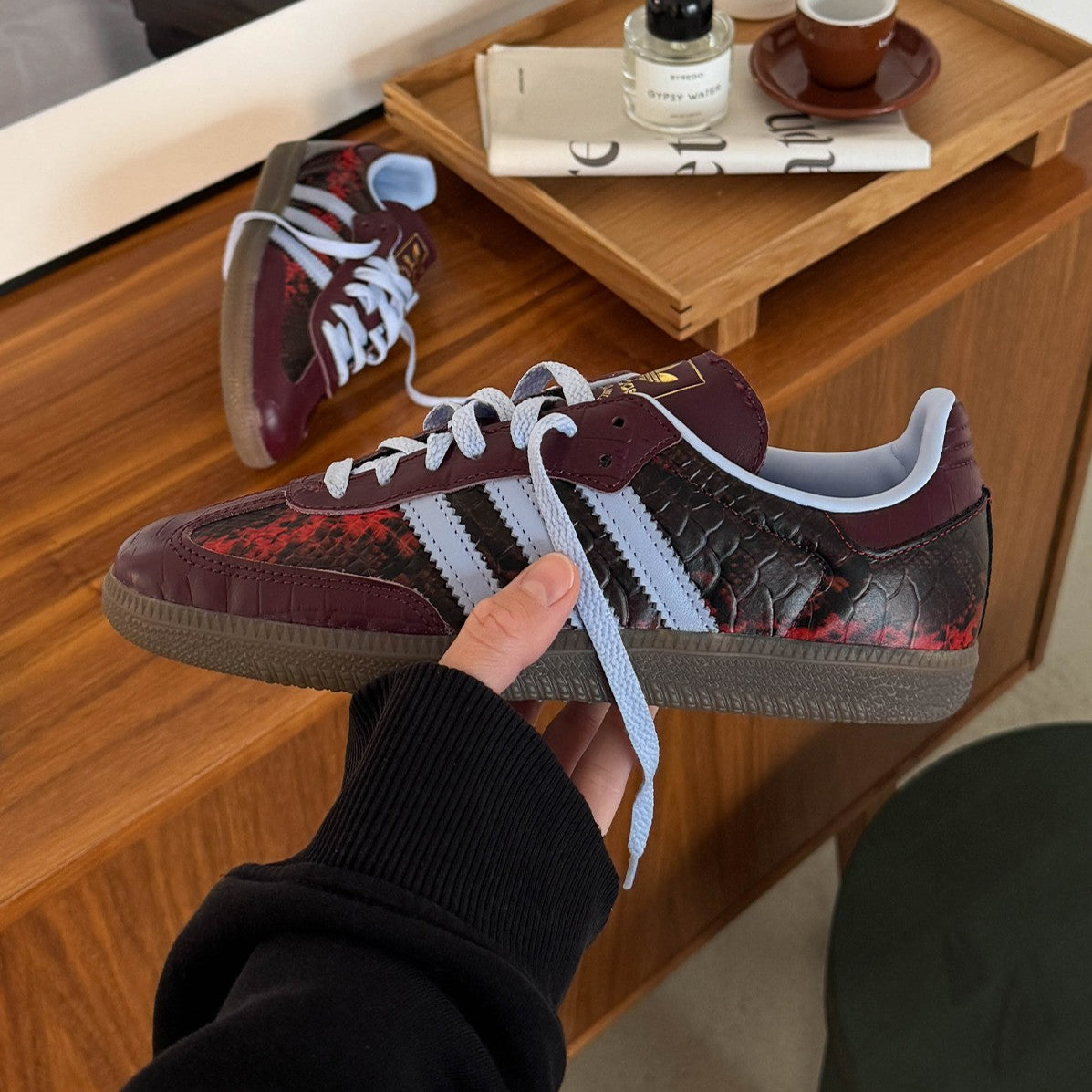 Hand hält einen bordeauxroten Terrace-Sneaker mit hellblauen Streifen über einen Holztisch