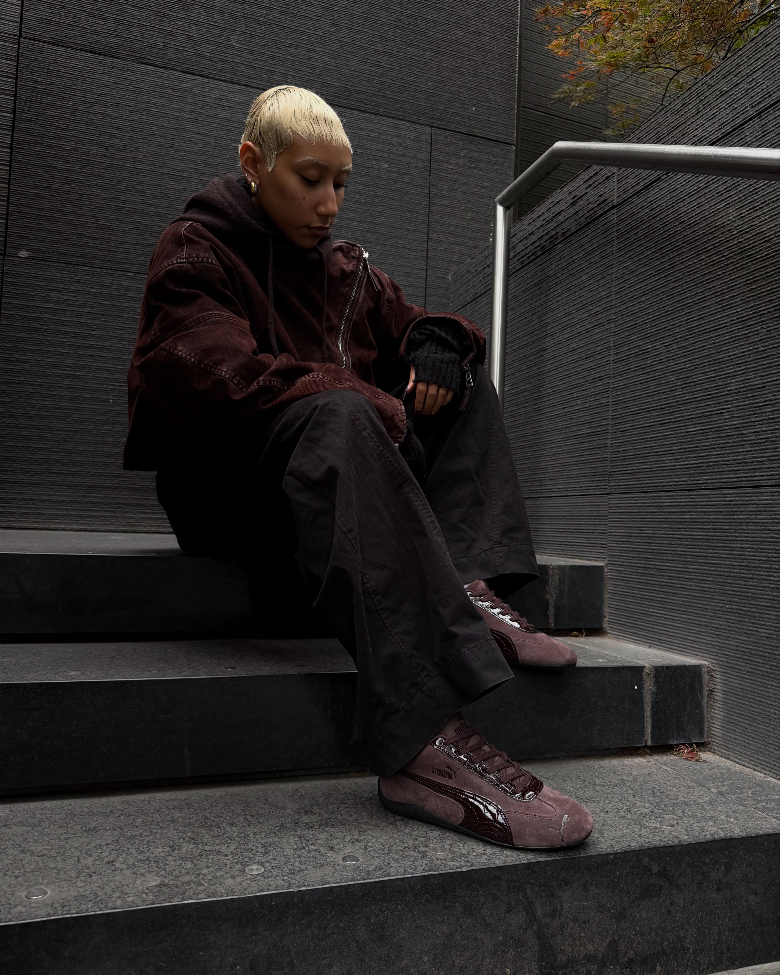 Model in a brown suede jacket and dark wide trousers sitting on stone steps wearing the PUMA Speedcat Croco in Chocolate Fondue