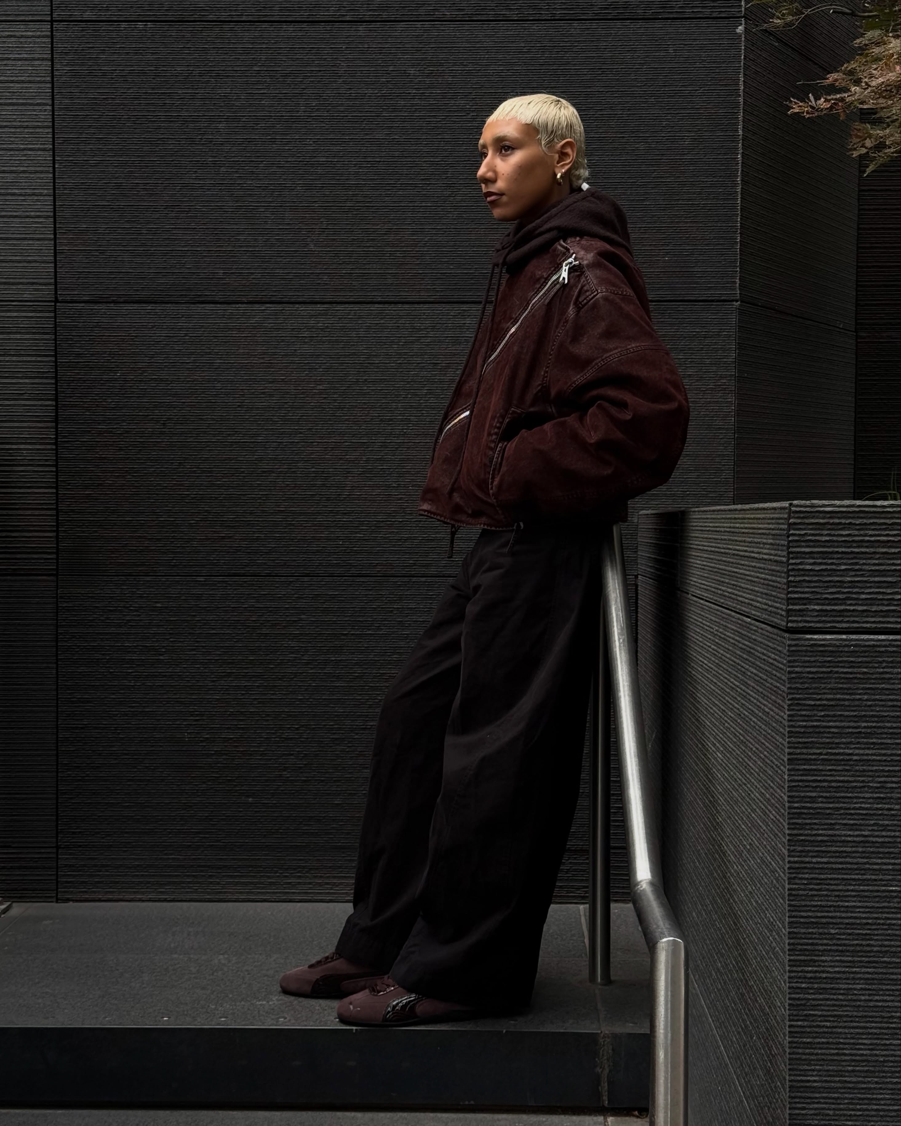 Model in a dark brown jacket and wide trousers leaning on a railing, wearing the PUMA Speedcat Croco in Chocolate Fondue