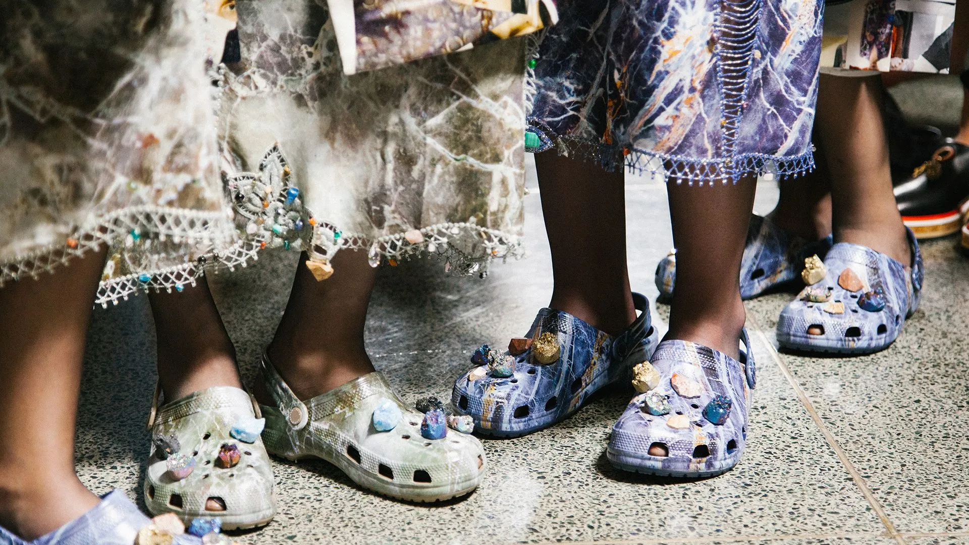Close-up of designer Crocs worn with embellished fashion looks on a runway, showcasing beaded and stone-decorated Jibbitz charms.