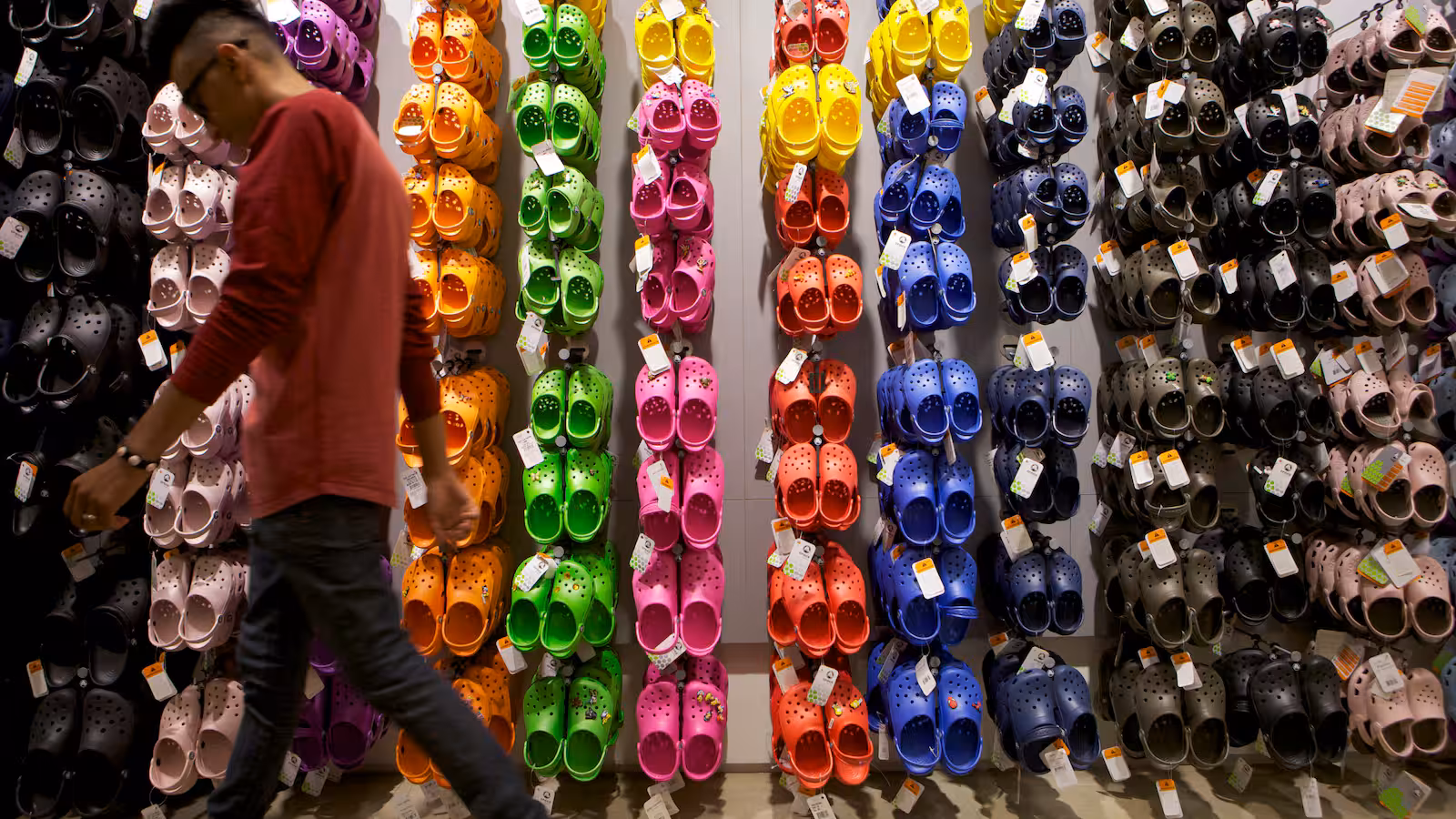 Person walking past a colorful wall display of Crocs in a retail store, showcasing a wide range of Crocs styles and colors from black to neon green.