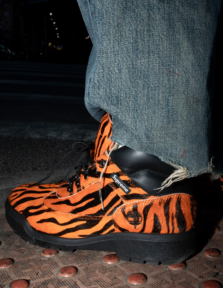 Close-up of an orange-and-black tiger-striped Timberland boot with a Supreme label, worn with wide denim jeans on a textured metal surface.
