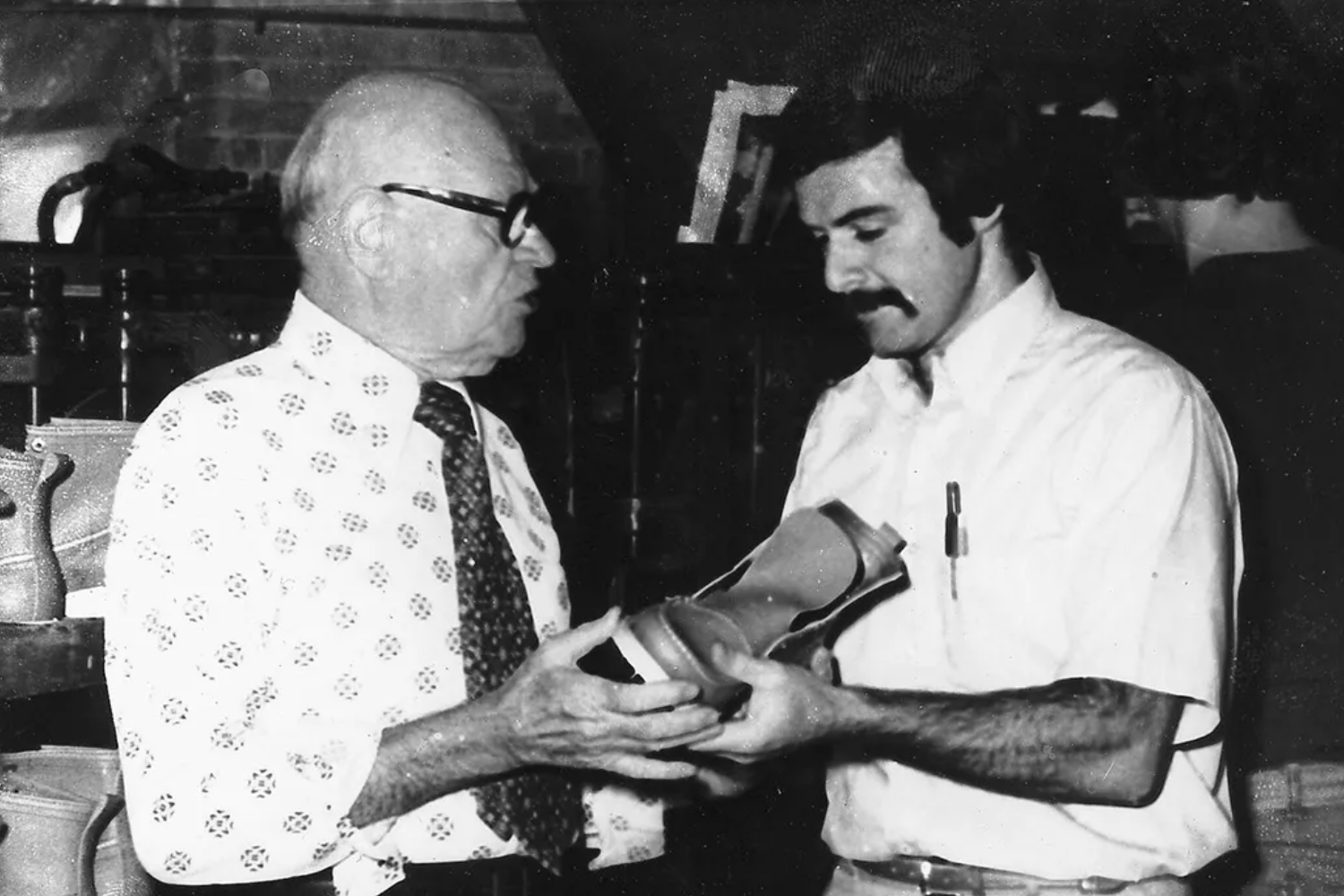 Black-and-white photograph of Nathan Swartz with his son, Sidney Swartz examining a shoe prototype, representing traditional footwear craftsmanship.