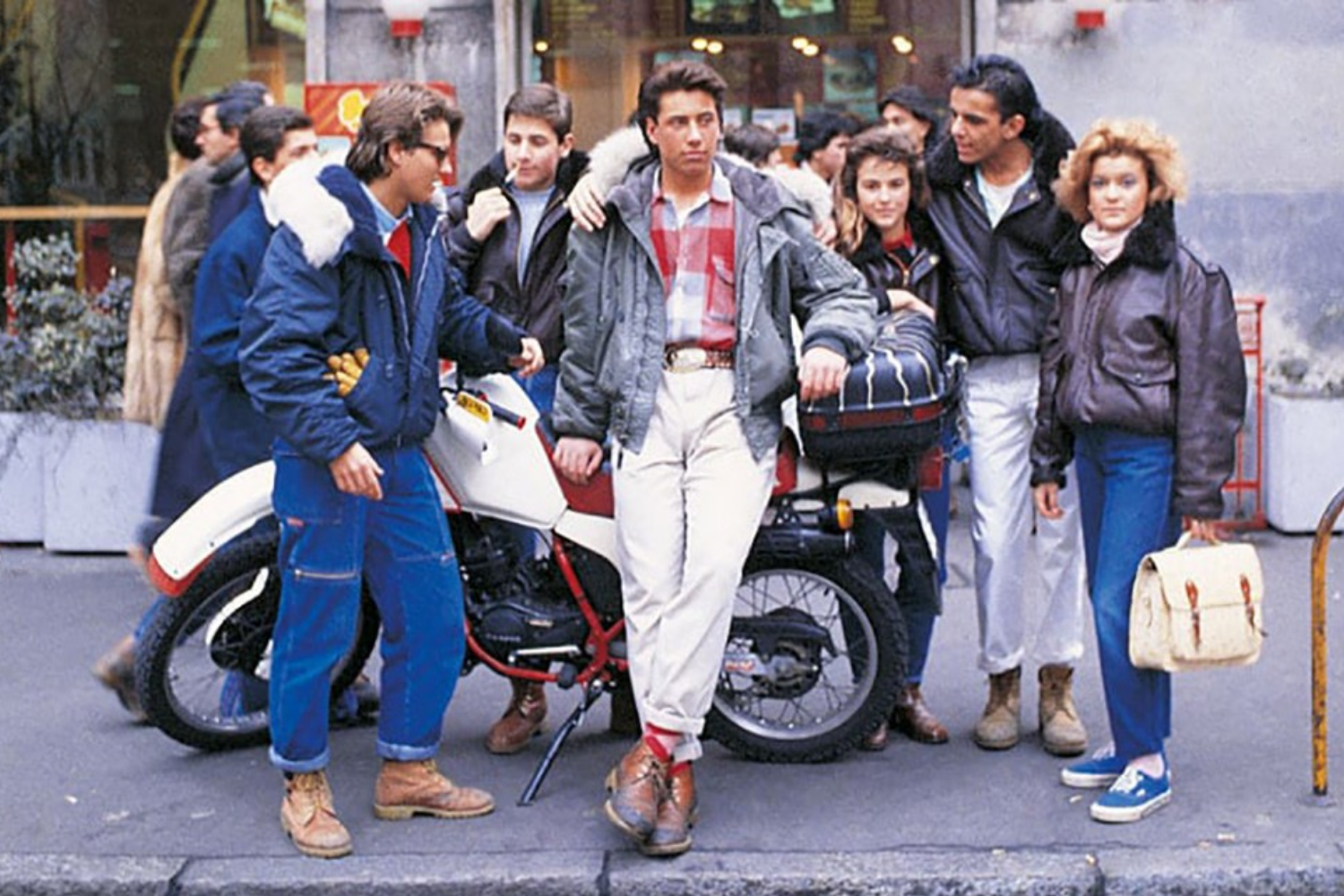 Group of young adults gathered around a motorcycle wearing casual jackets and boots, reflecting European street fashion of the 1980s.