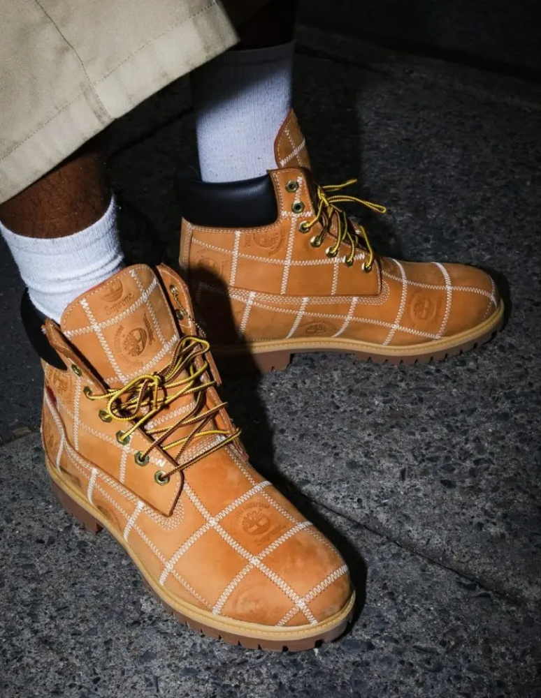Close-up of tan Timberland boots with stitched detailing, worn with white socks on a city sidewalk, highlighting the rugged sole and leather texture.