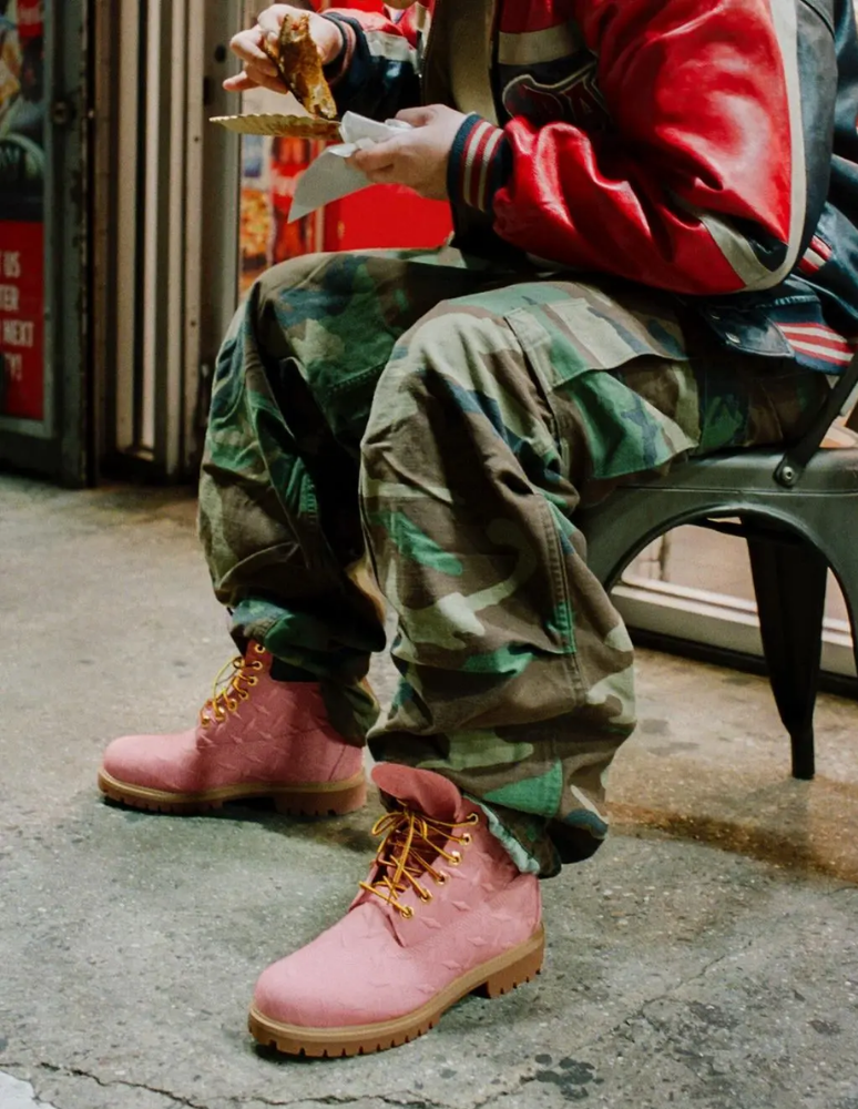 Person sitting on a sidewalk wearing pink Timberland boots with yellow laces, camouflage cargo pants, and a red jacket, eating street food in an urban setting.