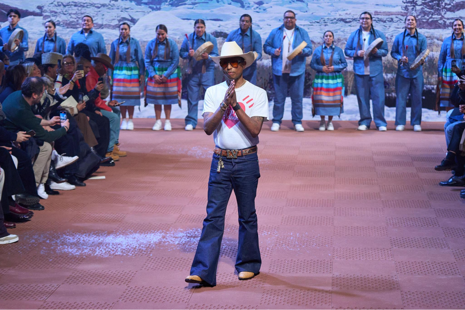 Pharrell Williams standing at the end of a runway wearing a white cowboy hat, jeans, and Timberland boots, with a group of performers in coordinated blue outfits behind him.